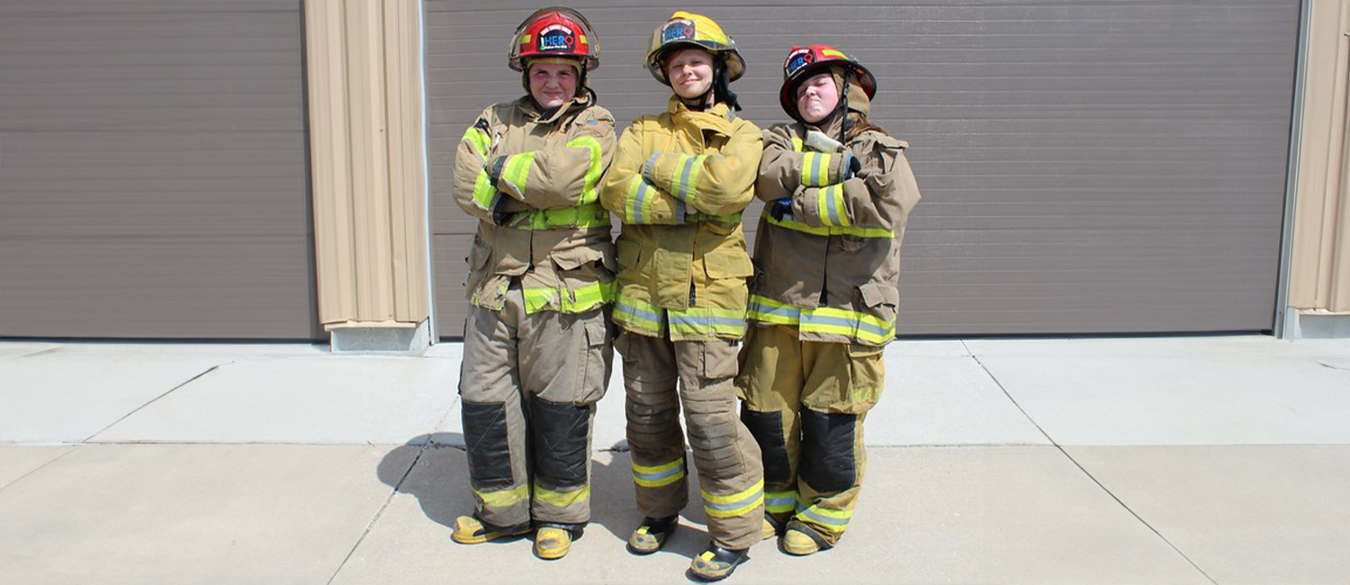 three teenage girls, dressed in firefighter uniforms and helmets, standing with crossed arms