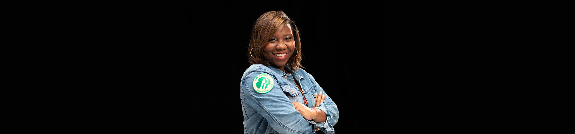  High school senior ambassador Girl Scout standing against black background and smiling at the camera - wearing a denim jacket with vintage Girl Scout profiles patch. 