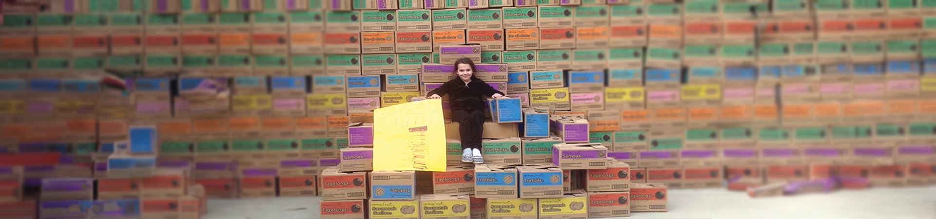  A young girl sitting on a throne made of Girl Scout Cookie cases in front of a wall of cookie cases 